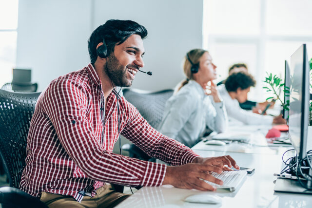 Smiling handsome ethnicity businessman working in call center. Shot of a cheerful young man working in a call center with his team. Confident male operator is working with colleagues. Call center operators sitting in a row at desks.