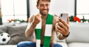 Young hispanic man supporting soccer match using smartphone sitting by christmas decor at home