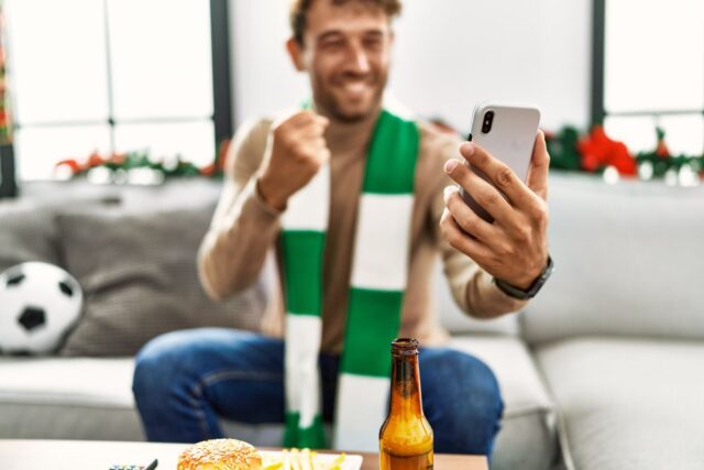 Young hispanic man supporting soccer match using smartphone sitting by christmas decor at home